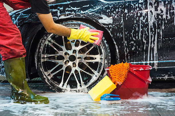 Person washing a car wheel with soapy water, wearing gloves and green rubber boots.